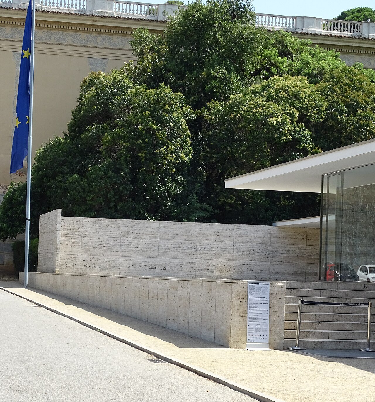 The corner of the pavilion's travertine wall and flat roof, beside a tree and a flagpole.