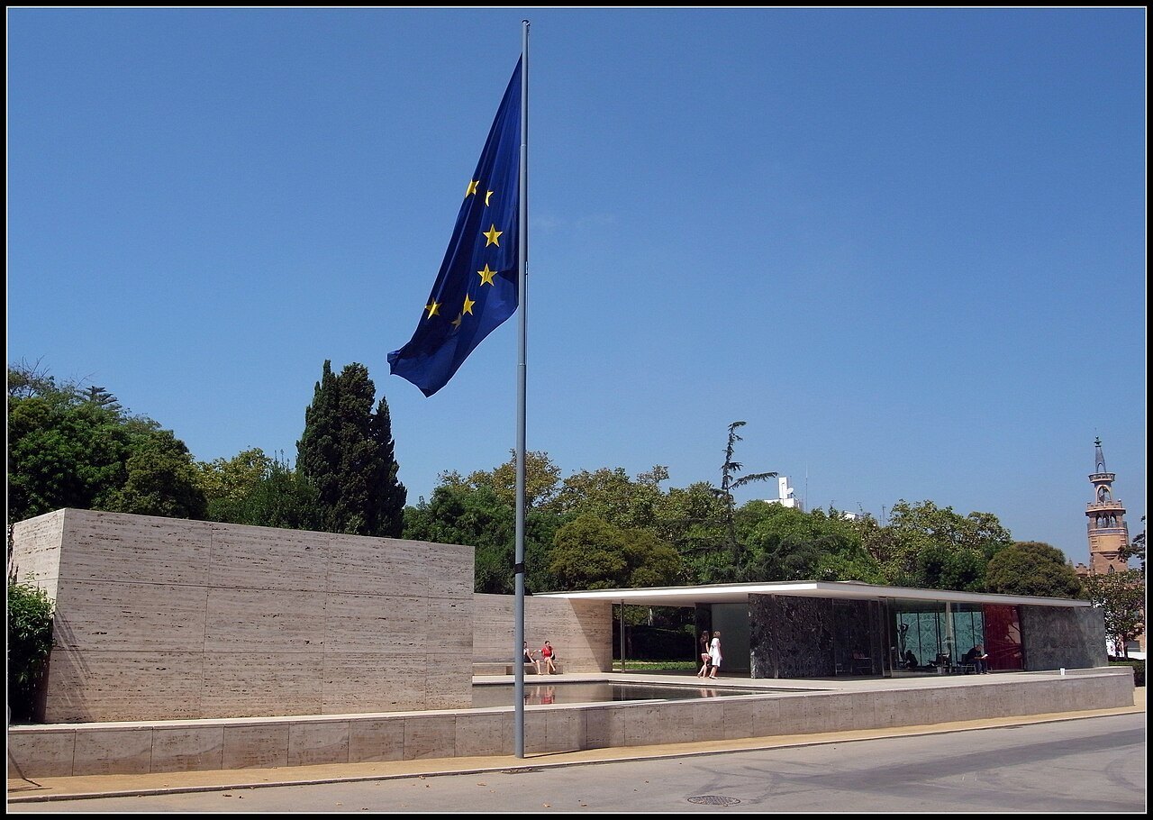The pavilion from across the street, a low travertine wall and glass behind a flagpole.