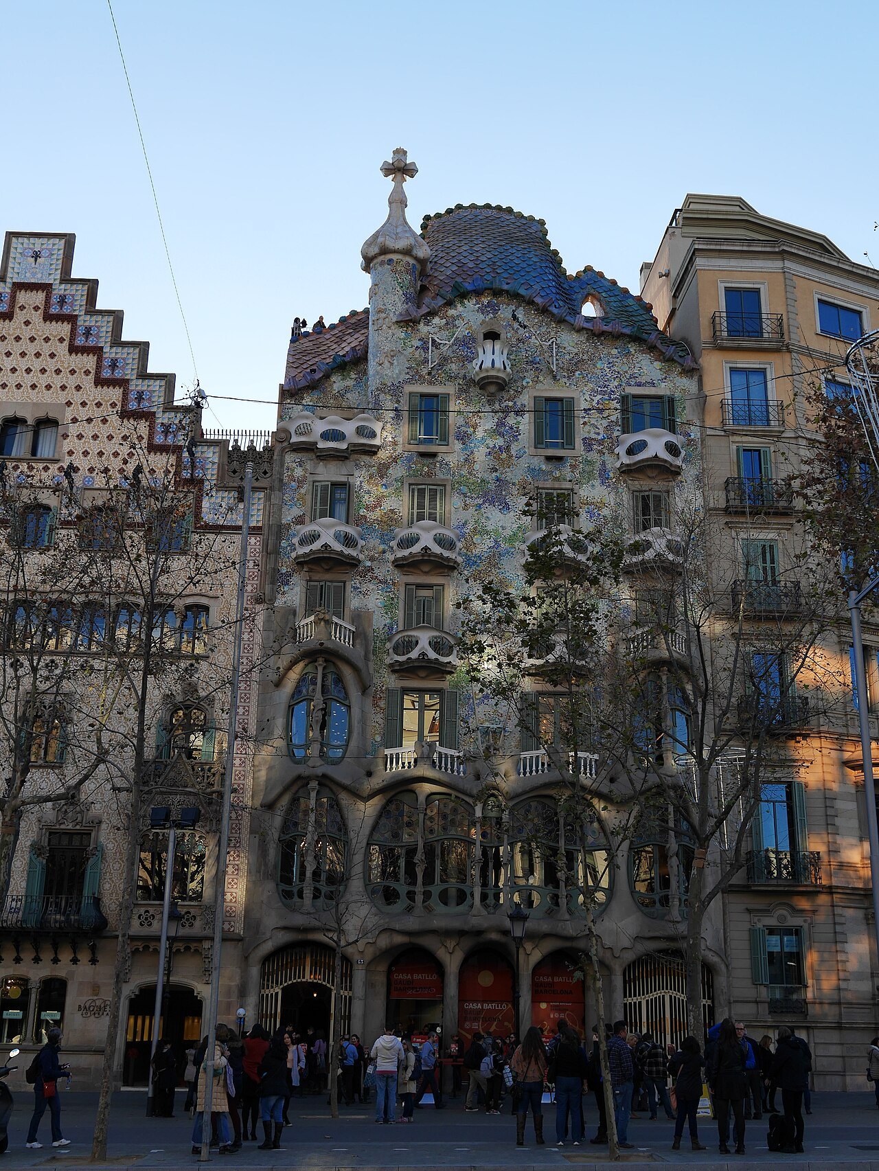 The front of Casa Batlló in daylight, with the stepped gable of Casa Amatller at left and visitors gathered in the street.