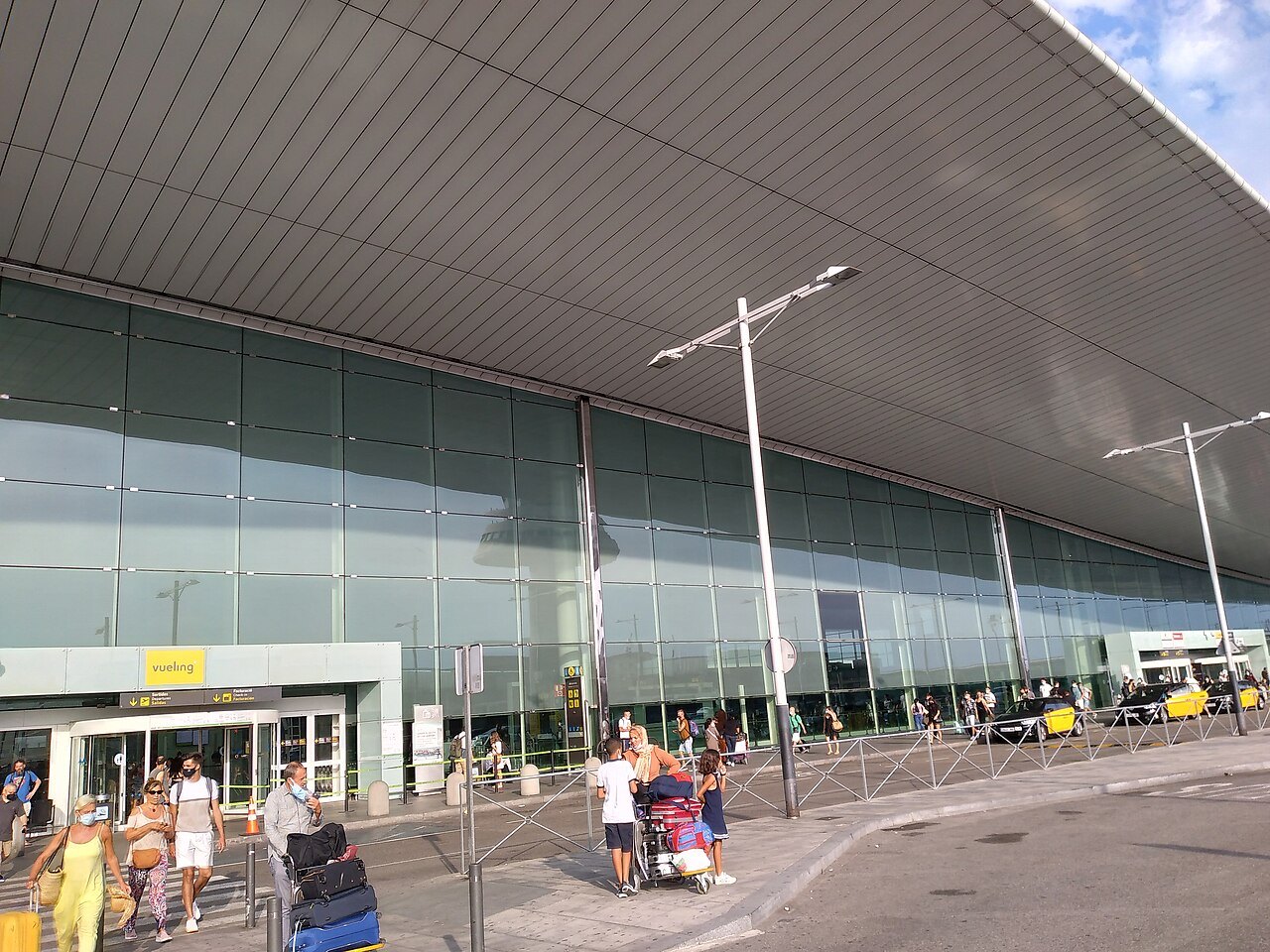 The front of an airport terminal, with arriving travelers and a taxi lane.