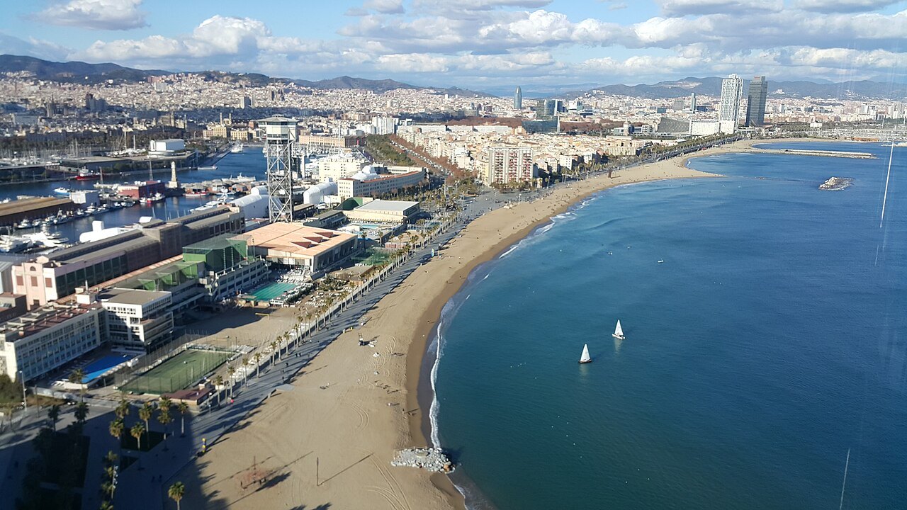 The curve of the Sant Sebastià and Barceloneta beach from above, with sailboats offshore.