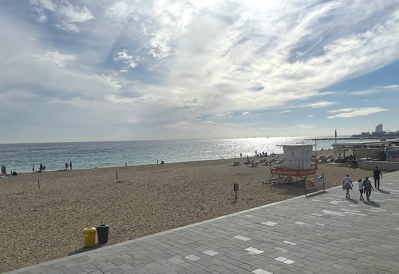 A wide, quiet beach at Bogatell, with a paved promenade and broken cloud overhead.