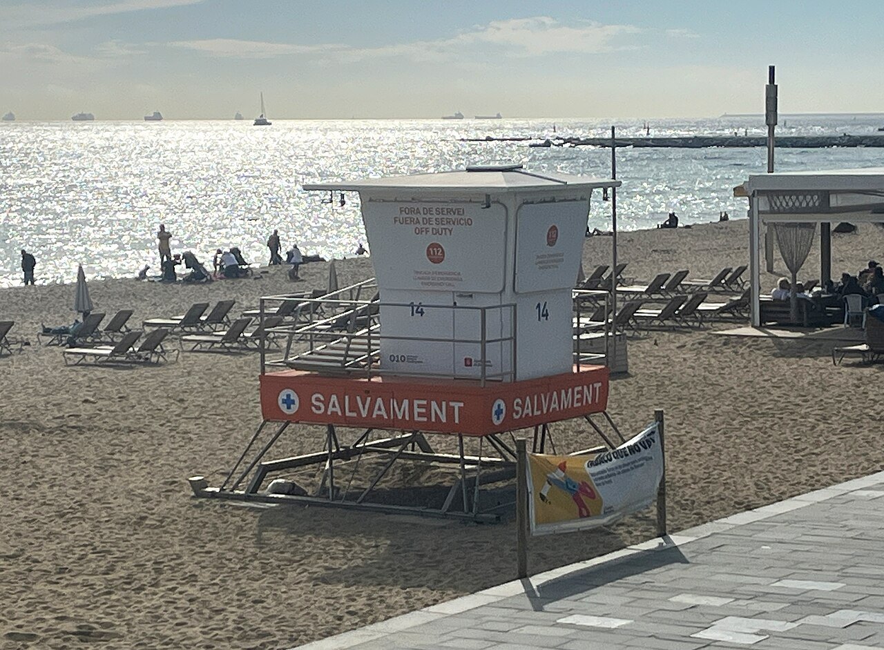 A white lifeguard tower on the sand at Bogatell, with sunbeds beyond.