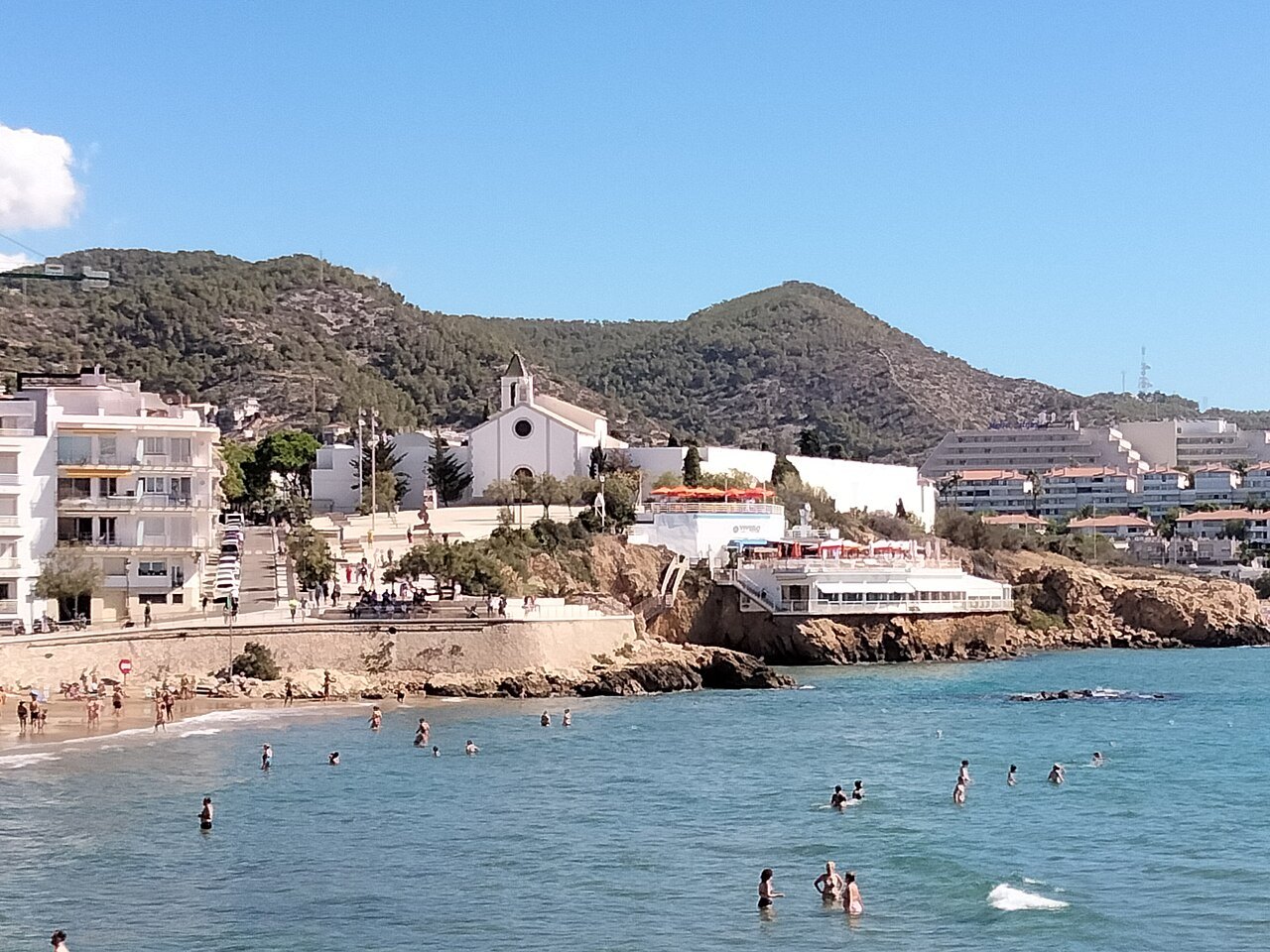 The beach at Sitges below a small church on the headland, with hills behind.