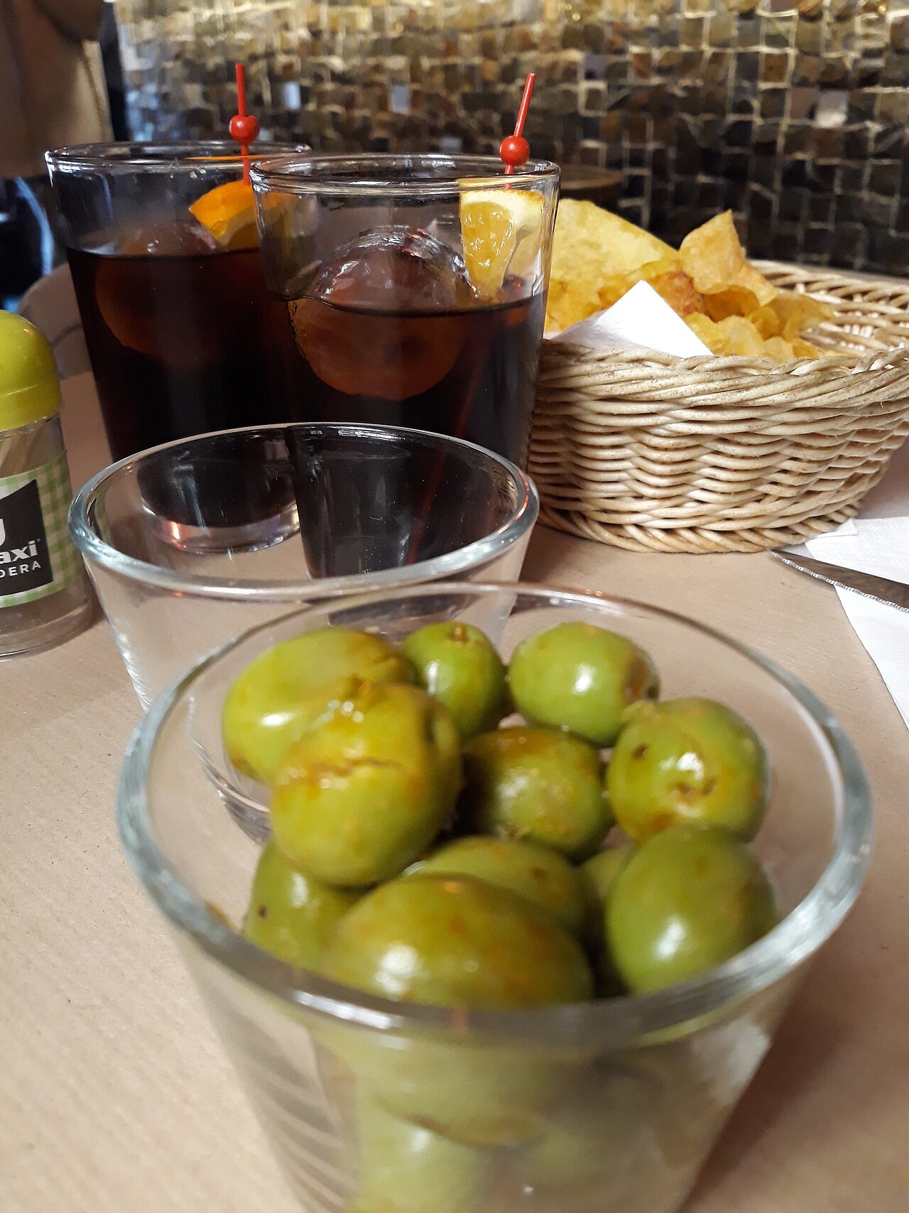 Two glasses of vermouth with a glass of green olives and a basket of chips.