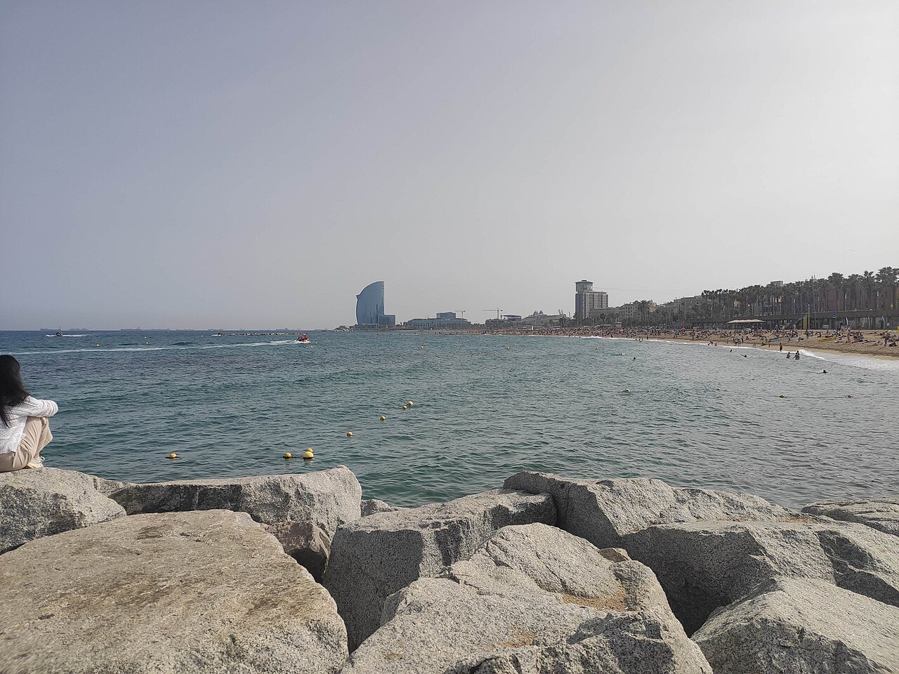 Breakwater rocks in front of a hazy beach, with the hotel tower on the skyline.