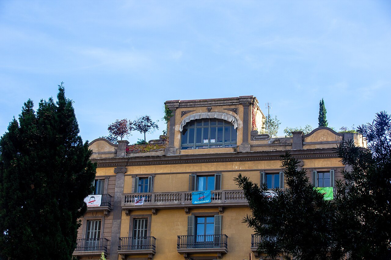 An ochre apartment building with plants on its roof and banners on its balconies, seen over trees.
