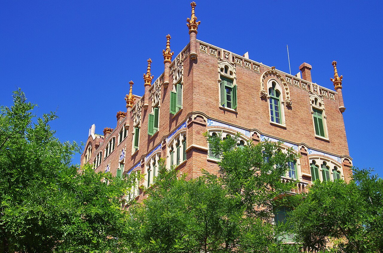 A brick pavilion with towers and green shutters, seen through trees at Sant Pau.