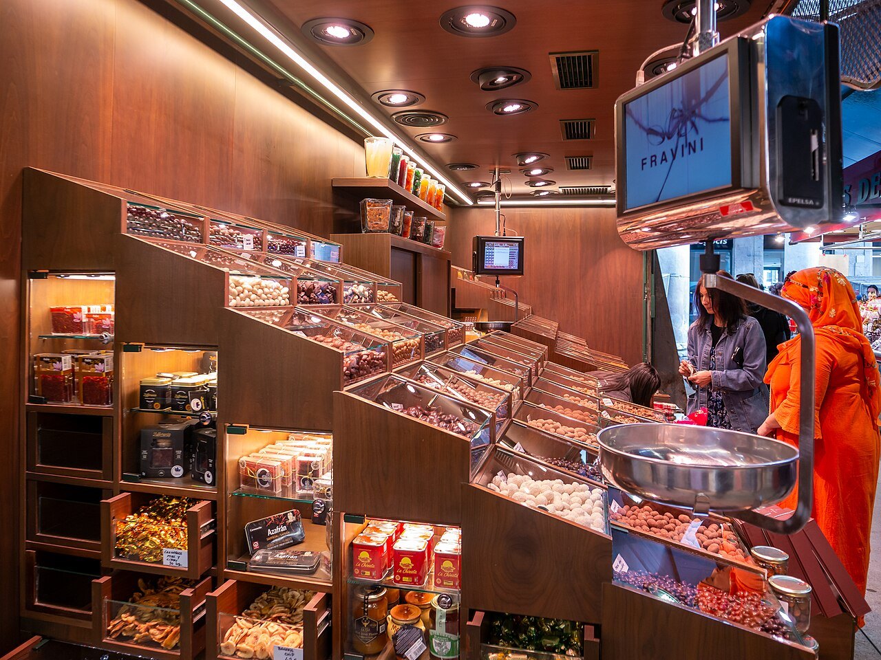 A stall of dried fruit and nuts on wooden shelving in the Boqueria, with a shopper in an orange sari.