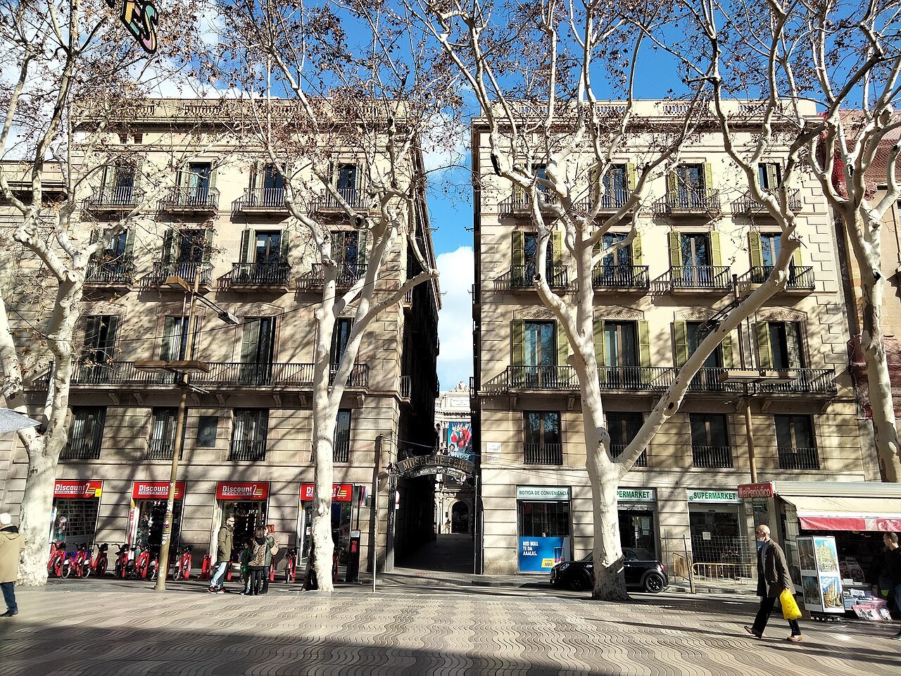 Plane trees and apartment blocks on the Rambla, with a narrow passage between two buildings.