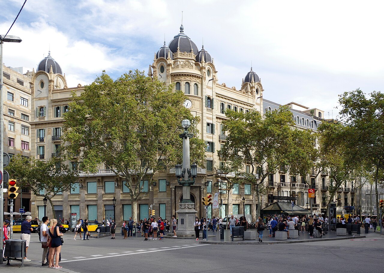 A domed corner building across a tree-lined square on the Rambla, with people crossing the street.