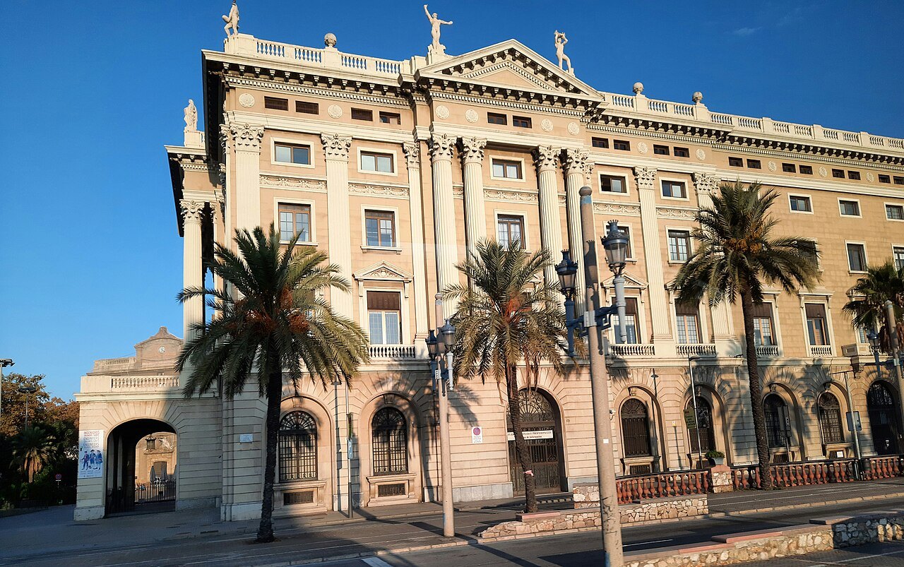 A grand stone building behind palm trees and street lamps, seen from the street.