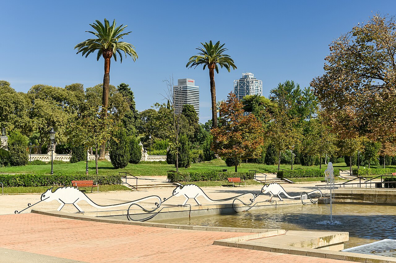 A pond with outlined lizard sculptures in the Parc de la Ciutadella, palms and tall towers behind.