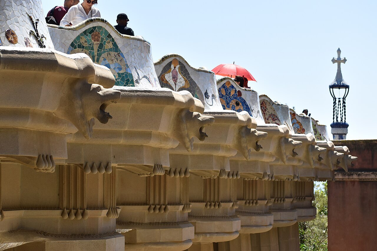 The serpentine bench at Park Güell, its mosaic-covered back running along the terrace, with a lamp topped by a cross.