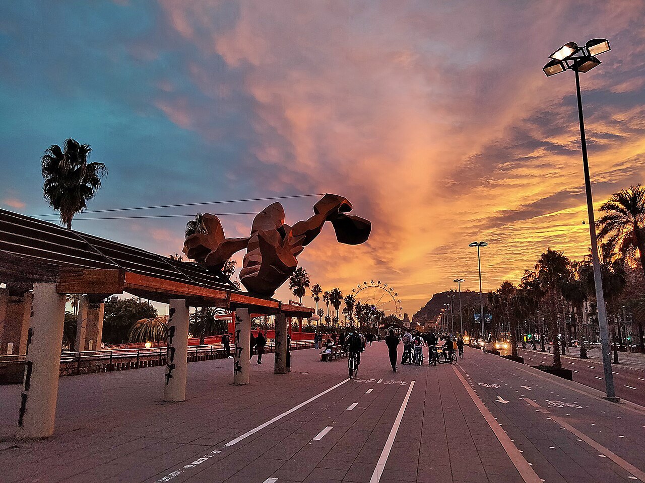 The harbor promenade at sunset under pink clouds, with a large sculpture beside it.