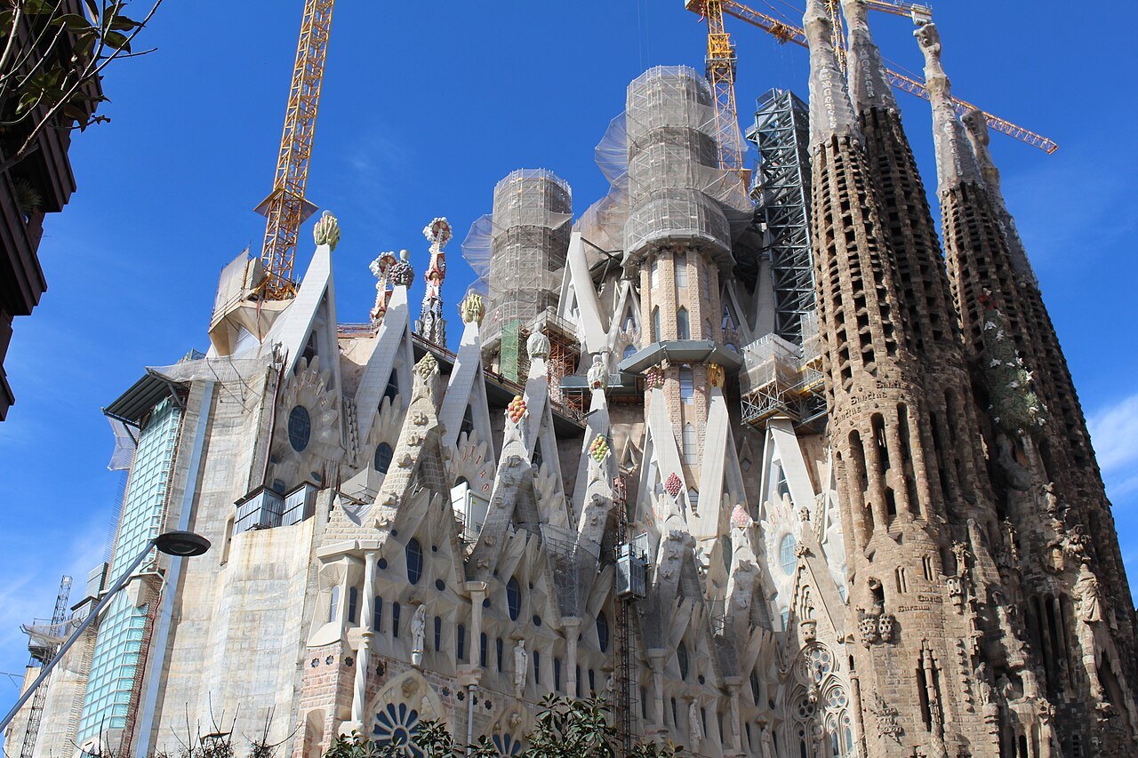 Cranes and scaffolding above the pale spires of the basilica, under a deep blue sky.