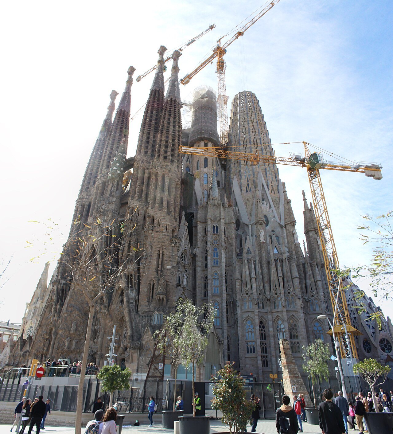 The basilica's towers behind cranes, with visitors in the street below.