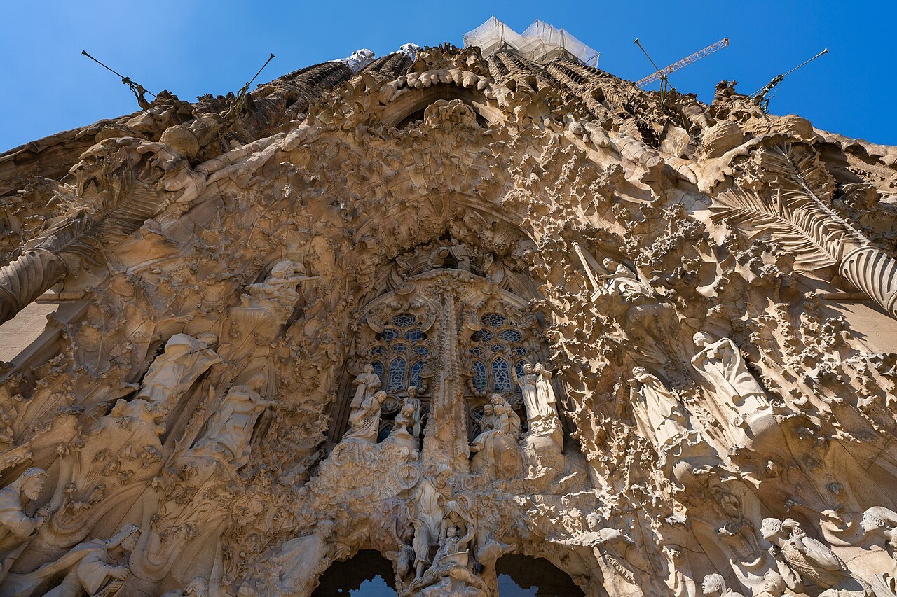 The carved stone of the Nativity façade of the Sagrada Família seen from below, with cranes at the top.