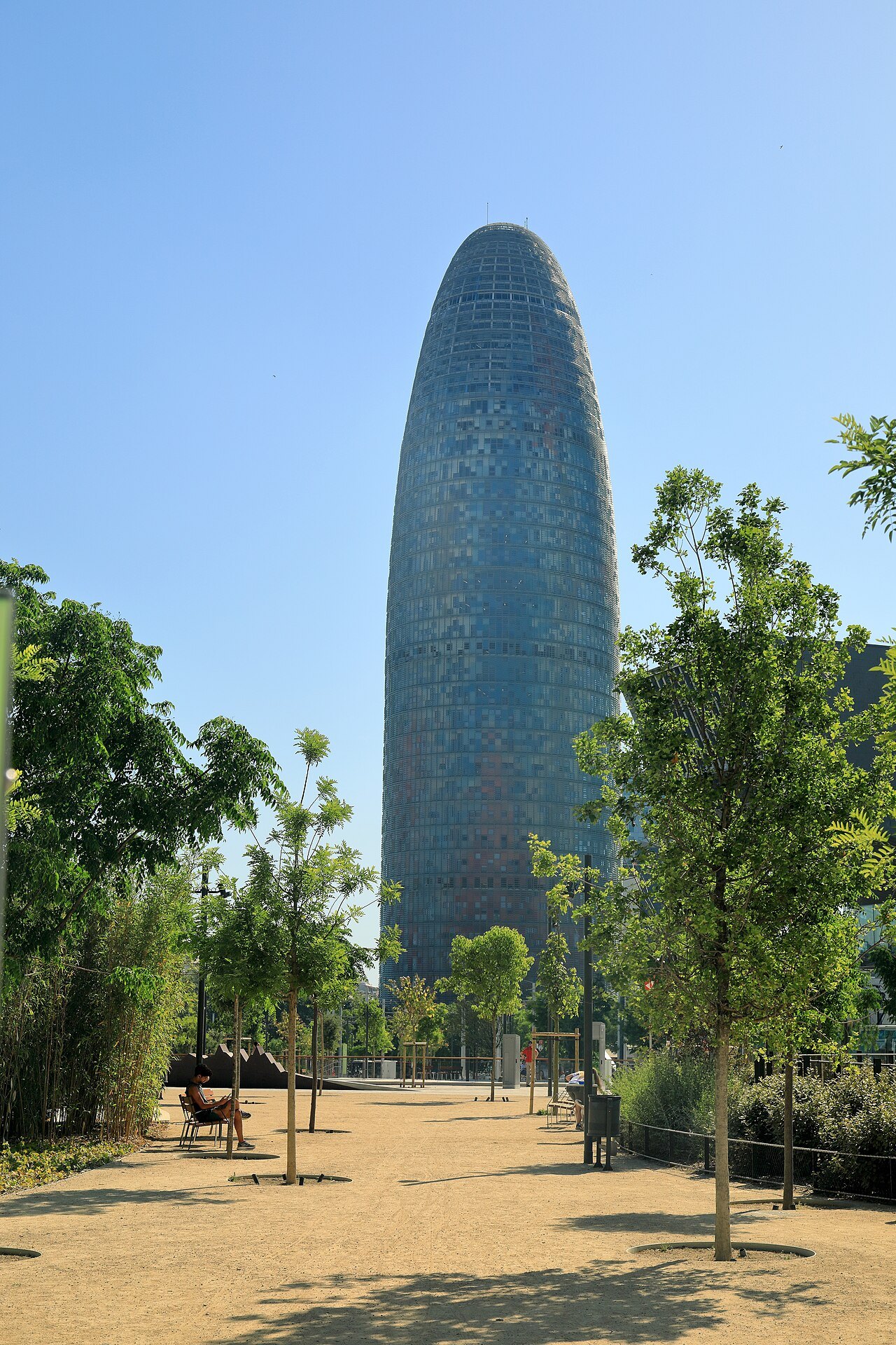The Torre Glòries rising above a park path and young trees.